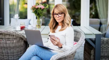 Eine blonde Frau mit Brille sitzt auf ihrer Terrasse und schaut auf ihren Laptop und ihr Smartphone.