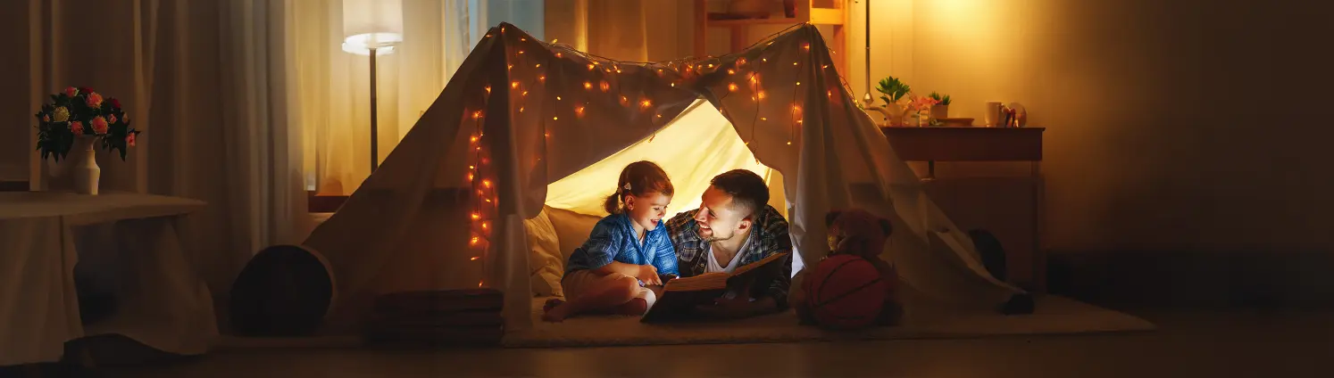Vater und Tochter kampieren abends beim Schein einer Lichterkette in einem selbst gebauten Zelt und lesen ein Buch.