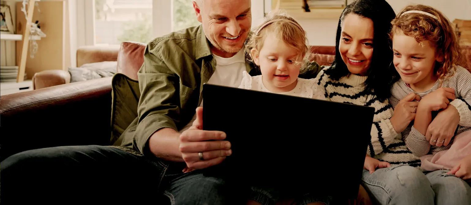 Vater Mutter und zwei Kinder sitzen zusammen auf dem Sofa und schauen lächelnd auf den Bildschirm eines Laptops.