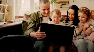 Vater Mutter und zwei Kinder sitzen zusammen auf dem Sofa und schauen lächelnd auf den Bildschirm eines Laptops.