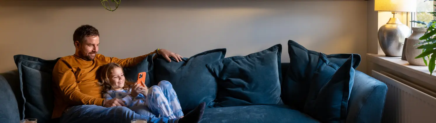 Ein Vater sitzt mit seiner Tochter gemütlich auf einem blaugrünen Sofa. Die Tochter lehnt sich an, während sie lachend auf ihr Mobiltelefon schaut.