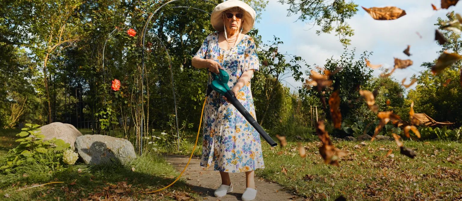Eine ältere Dame mit Sonnenhut und Sonnenbrille setzt elektrischen Laubbläser ein, um das Laub im Garten zu entfernen.