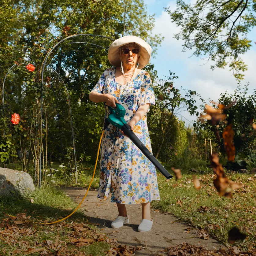 Eine ältere Dame mit Sonnenhut und Sonnenbrille setzt elektrischen Laubbläser ein, um das Laub im Garten zu entfernen.