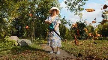 Eine ältere Dame mit Sonnenhut und Sonnenbrille setzt elektrischen Laubbläser ein, um das Laub im Garten zu entfernen.