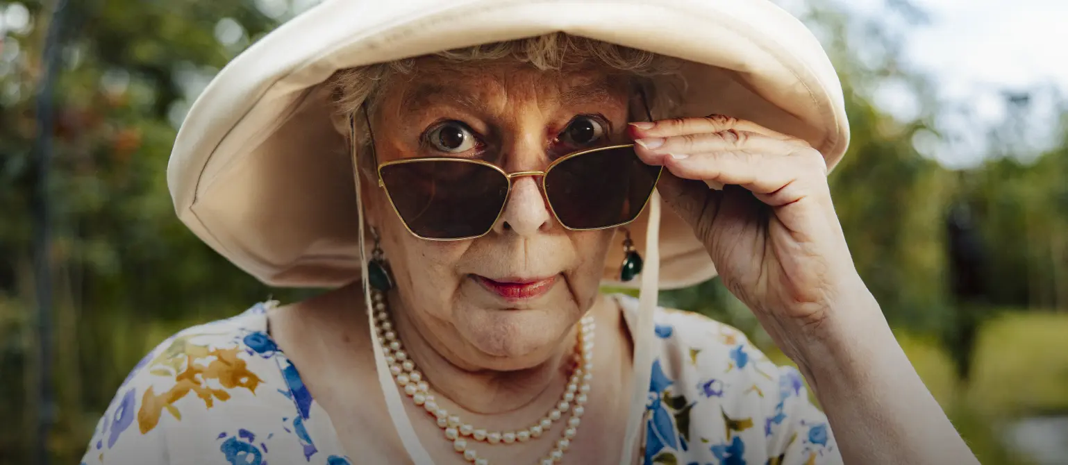 Ältere Frau schiebt ihre Sonnenbrille tiefer auf die Nase und schaut direkt in die Kamera.