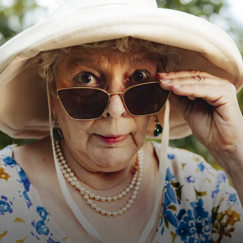 Ältere Frau schiebt ihre Sonnenbrille tiefer auf die Nase und schaut direkt in die Kamera.
