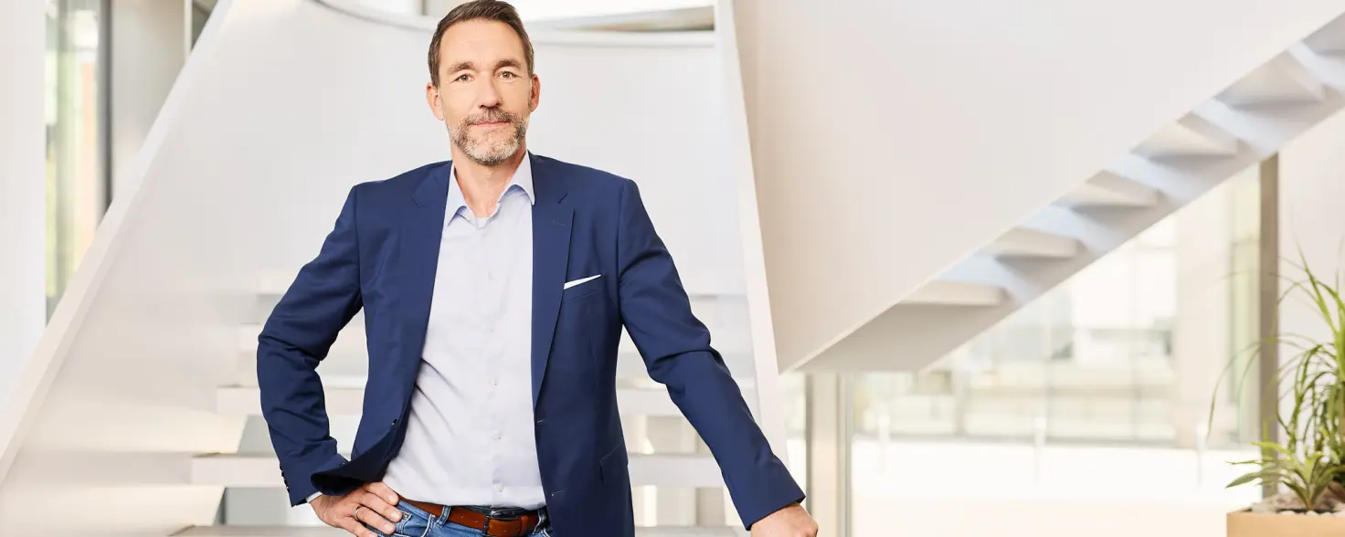 Ein Mitarbeiter von Energie Brandenburg in Sakko und Jeans steht in der Lobby vor einer weißen Treppe und lächelt.