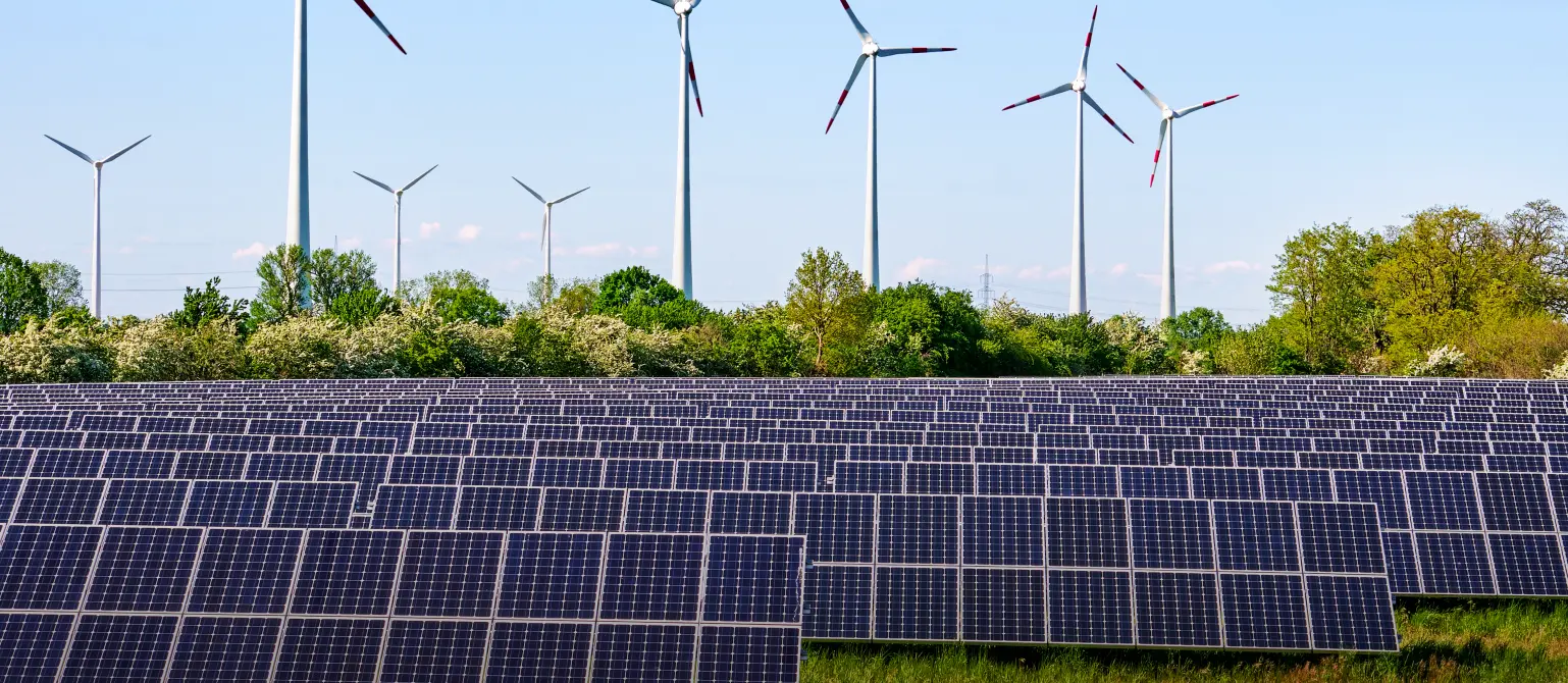 Ein Solarpark auf einer grünen Wiese. Dahinter ein kleiner Wald und acht Windkraftanlagen.