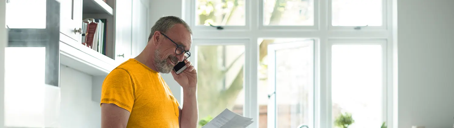 Ein Mann mit Brille lehnt an der Küchenzeile, er telefoniert während er einen geöffneten Brief hält.