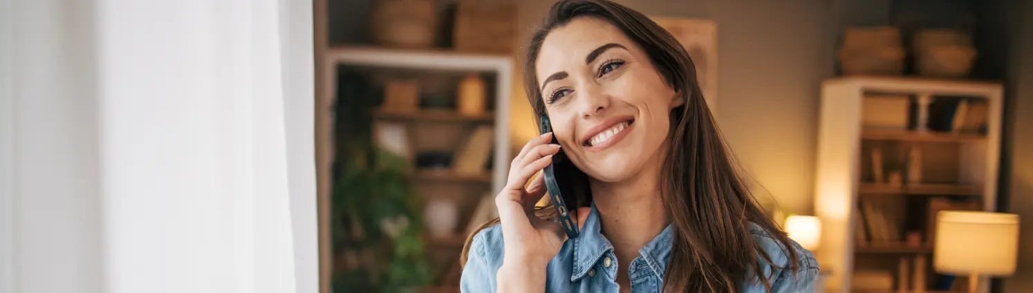 Lächelnde Frau im Jeanshemd steht im Wohnzimmer am Fenster und telefoniert mit dem Handy.