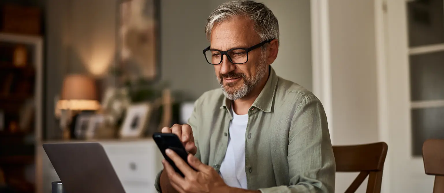 Ein lächelnder älterer Mann mit Brille, der ein Handy benutzt, während er an seinem Laptop arbeitet.