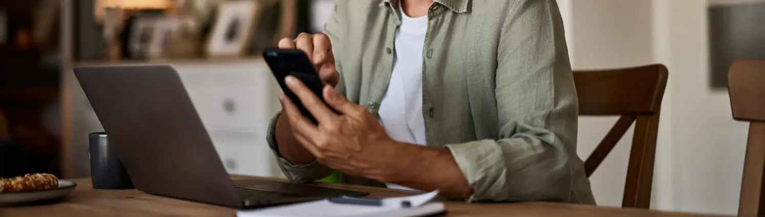 Ein lächelnder älterer Mann mit Brille, der ein Handy benutzt, während er an seinem Laptop arbeitet.