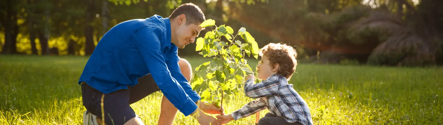 Ein Mann und sein Sohn knien am Boden und pflanzen einen Baum auf einer grünen Wiese.