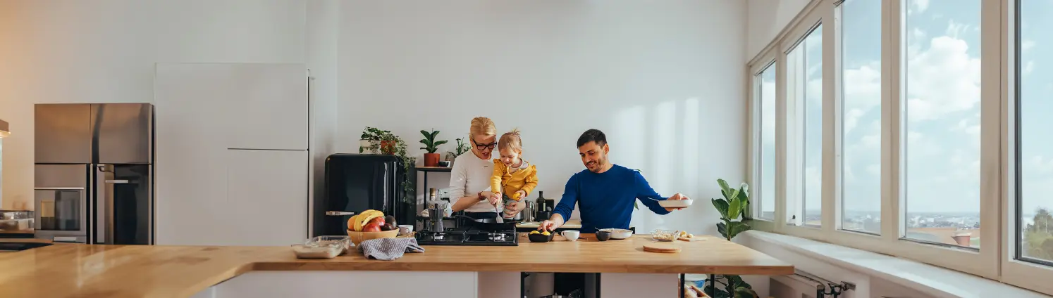 Eine Familie kocht in ihrer Küche. Der Vater stellt die Zutaten zusammen, während die Mutter mit dem Kind in der Pfanne auf dem Gasherd rührt.