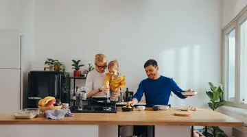 Eine Familie kocht in ihrer Küche. Der Vater stellt die Zutaten zusammen, während die Mutter mit dem Kind in der Pfanne auf dem Gasherd rührt.