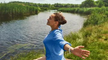 Eine lächelnde Frau im blau gestreiften Hemd kniet mit weit geöffneten Armen auf einem Steg am Fluss.