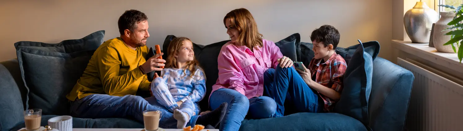 Eine vierköpfige Familie sitzt auf dem Sofa und trinkt Tee. Der Vater zeigt seiner Frau etwas auf dem Smartphone. Die Kinder lächeln.