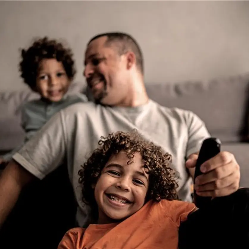 Ein lächelnder Jungen, der mit seinem Vater und seinem Bruder zu Hause auf dem Sofa fernsieht, hält eine Fernbedienung.