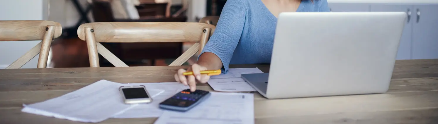 Einer junge Frau mit dunklem Haar, hellblauem Pulli und Brille sitzt in ihrer Küche vor dem Laptop und bedient einen Taschenrechner.