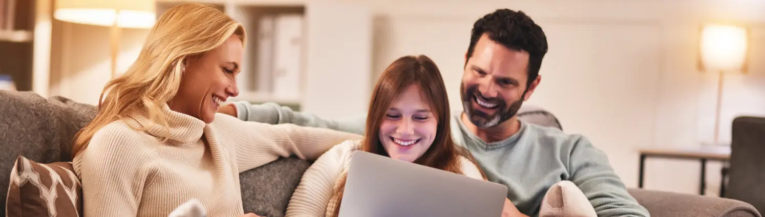 Vater, Mutter und Teenager-Tochter sitzen auf dem Sofa. Die Tochter hat die Beine hochgelegt und zeigt den Eltern etwas auf ihrem Laptop.