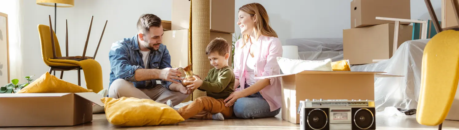 Vater, Kleinkind und Mutter sitzen in der neuen Wohnung auf dem Parkettboden, inmitten von Umzugskartons. Der Sohn betrachtet eine goldene Karaffe.