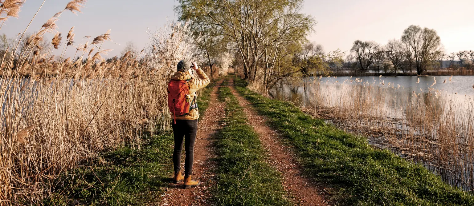 Eine junge Frau mit Rucksack steht auf einem Feldweg am See und beobachtet Wildvögel mit dem Fernglas.