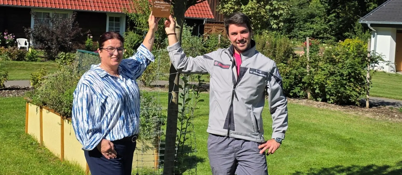 Garten des Kinderhauses Pusteblume: Eine Frau und ein Mann halten ein Schild, das an einem Baum befestigt ist und lächeln in die Kamera.