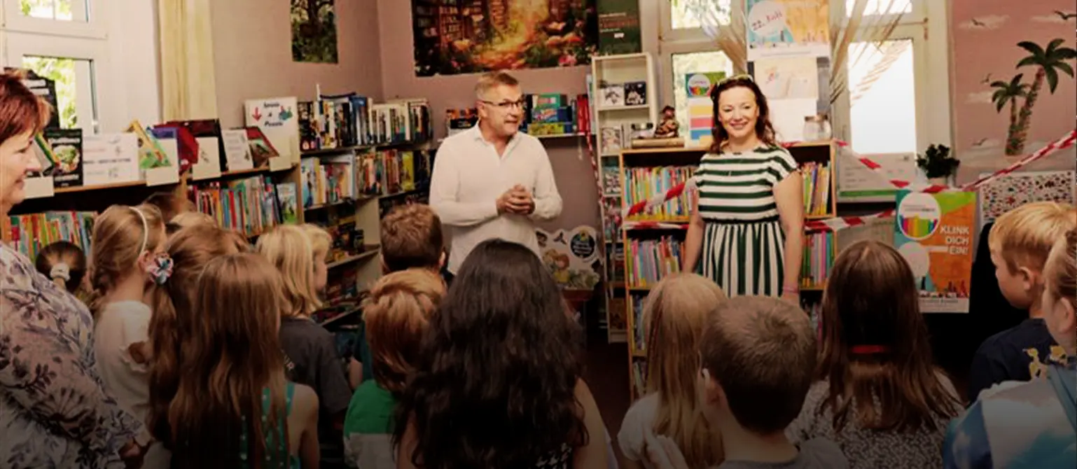 Eine Gruppe von Kindern lauscht in einer Bibliothek, die mit Luftballons geschmückt ist, beim Brandenburger Lesesommer den Veranstaltenden.