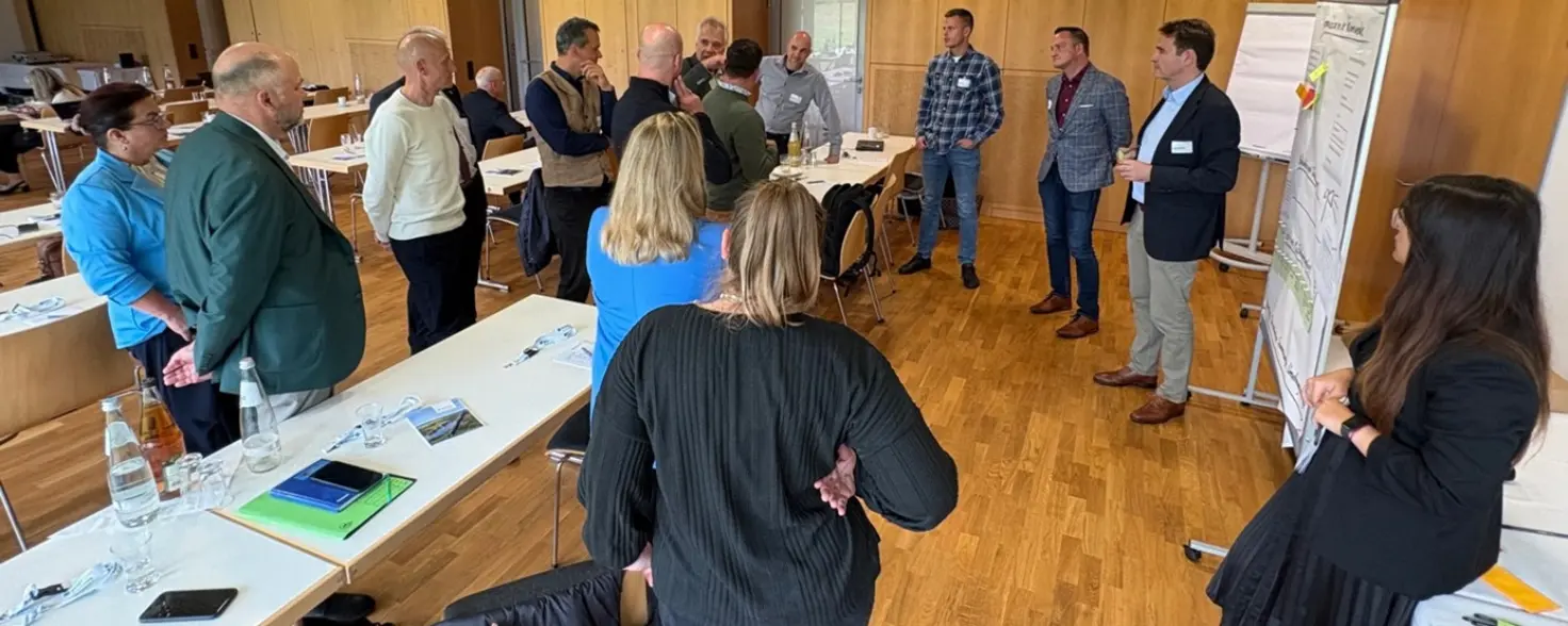 Gruppe von Personen steht in einem Seminarraum im Kreis und diskutiert vor Flipcharts. Im Hintergrund stehen Tische mit Unterlagen.