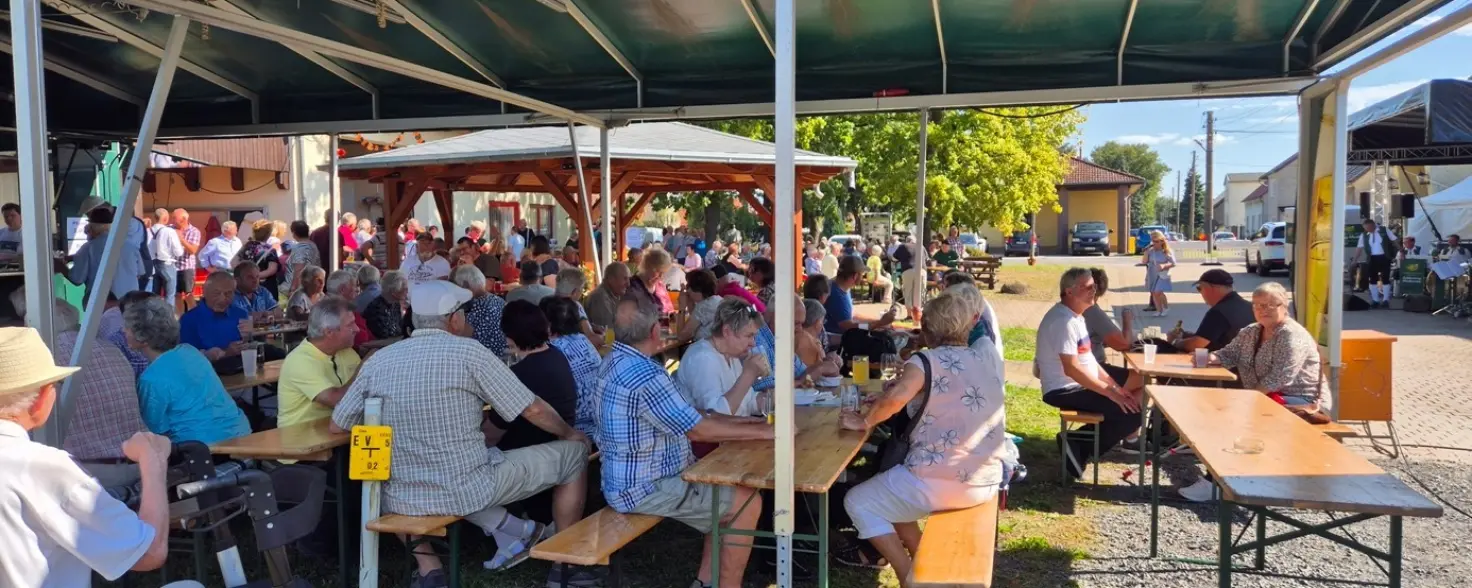 Vor einer Bühne sitzen unter einem Pavillon Menschen auf Bierbänken