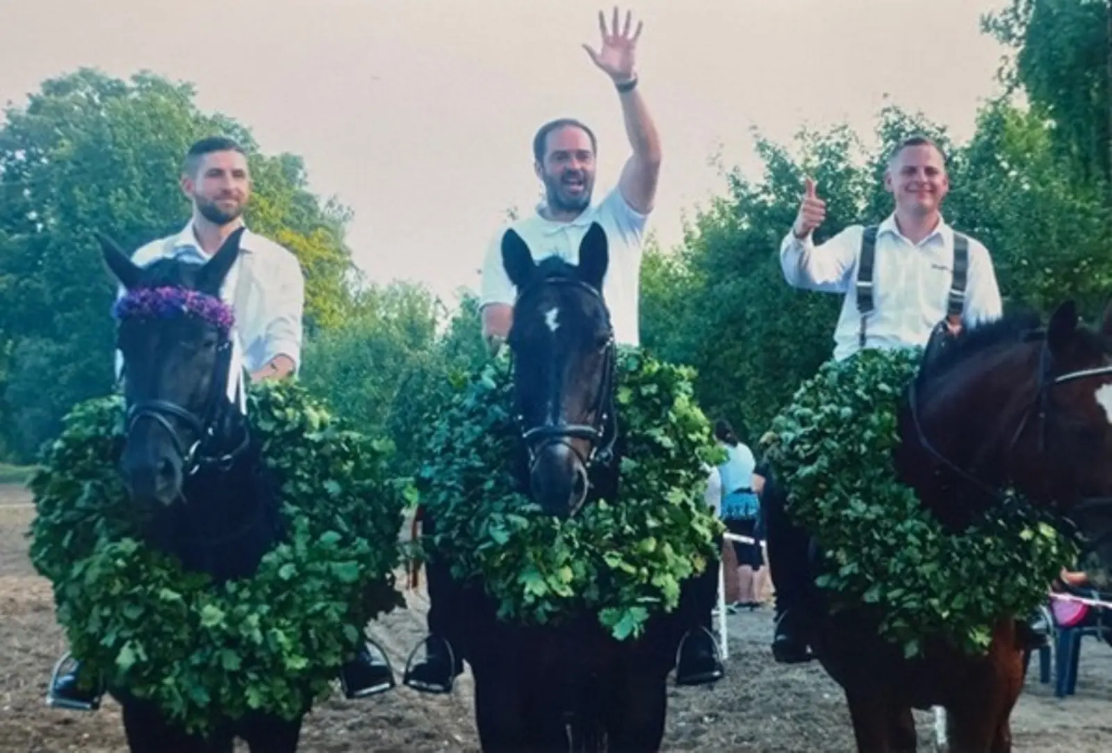 Drei Männer sitzen jeweils auf einem Pferd, der Mann in der Mitte winkt während der Mann auf der rechten Seite den Daumen nach oben zeigt