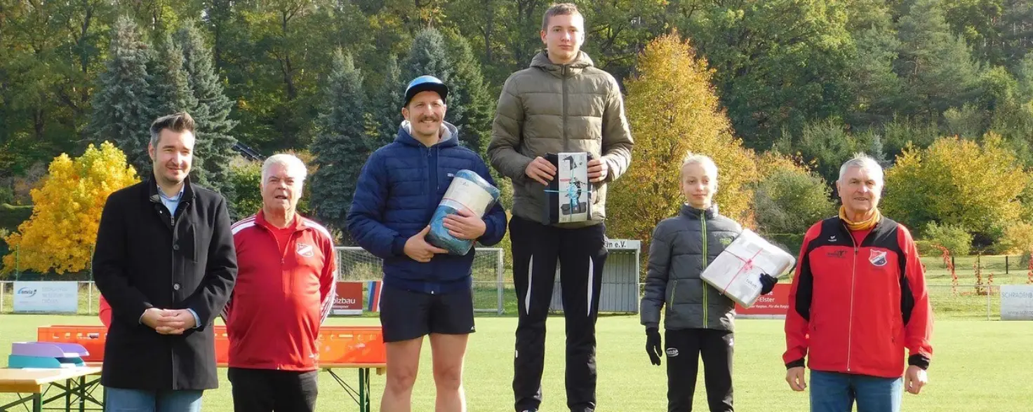 Drei Menschen stehen mit Urkunde in der Hand auf einer Tribüne, welche den Erst- bis Drittplatzierten zeigt