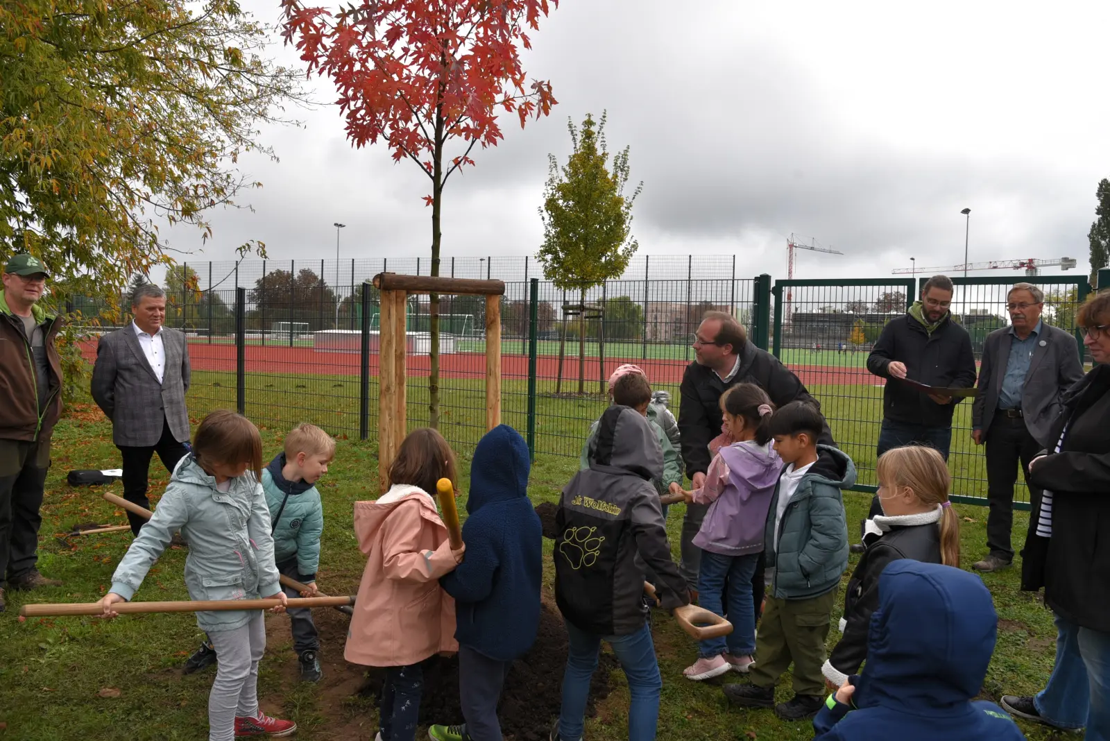 Neben einem Sportplatz buddeln Kinder gemeinsam ein Loch in die Erde und Erwachsene stehen drumherum
