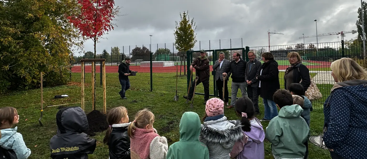 Eine Gruppe von Kindern und Erwachsenen stehen um einen frisch gepflanzten Baum drumherum