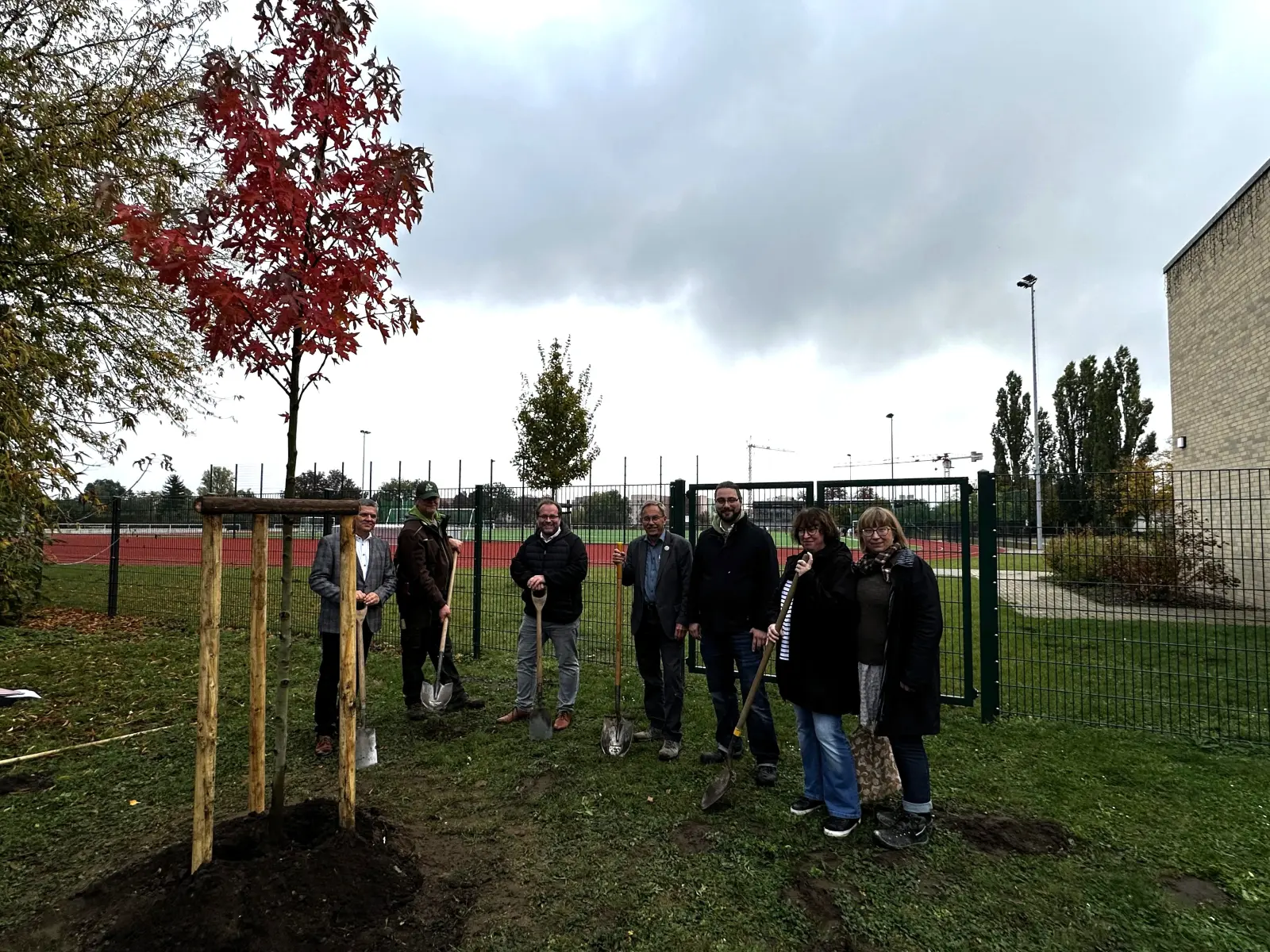 Erwachsene mit Schaufeln stehen hinter einen frisch gepflanzten Baum und lächeln in die Kamera