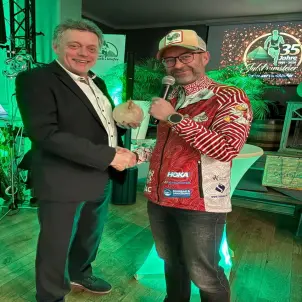 Zwei Männer reichen sich in einem grün beleuchteten Raum die Hand. Ein Mann mit Sportjacke hält ein Mikrofon, der Mann im Anzug ein Huhn aus Ton.