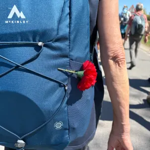 Eine rote Blume ist an einen dunkelblauen Rucksacks einer wandernden Person geklemmt.