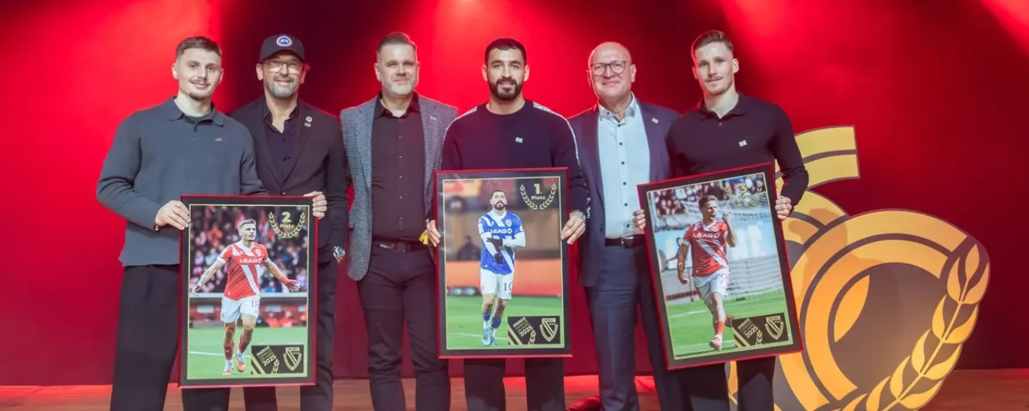 Sechs Männer stehen auf einer Bühne vor roter Beleuchtung. Drei von ihnen halten große Bilder von drei Fußballspielern in der Hand.