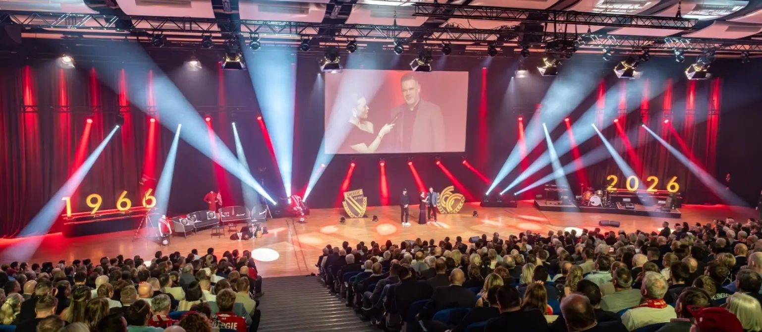 Eine Bühne mit einem großen Publikum. Durch den Saal schwenken rote und weiße Scheinwerfer.
