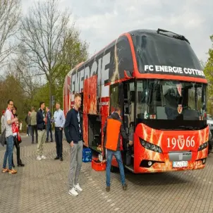 Ein roter Fan-Bus des FC Energie Cottbus steht auf einem Parkplatz. Einige Fans stehen davor.