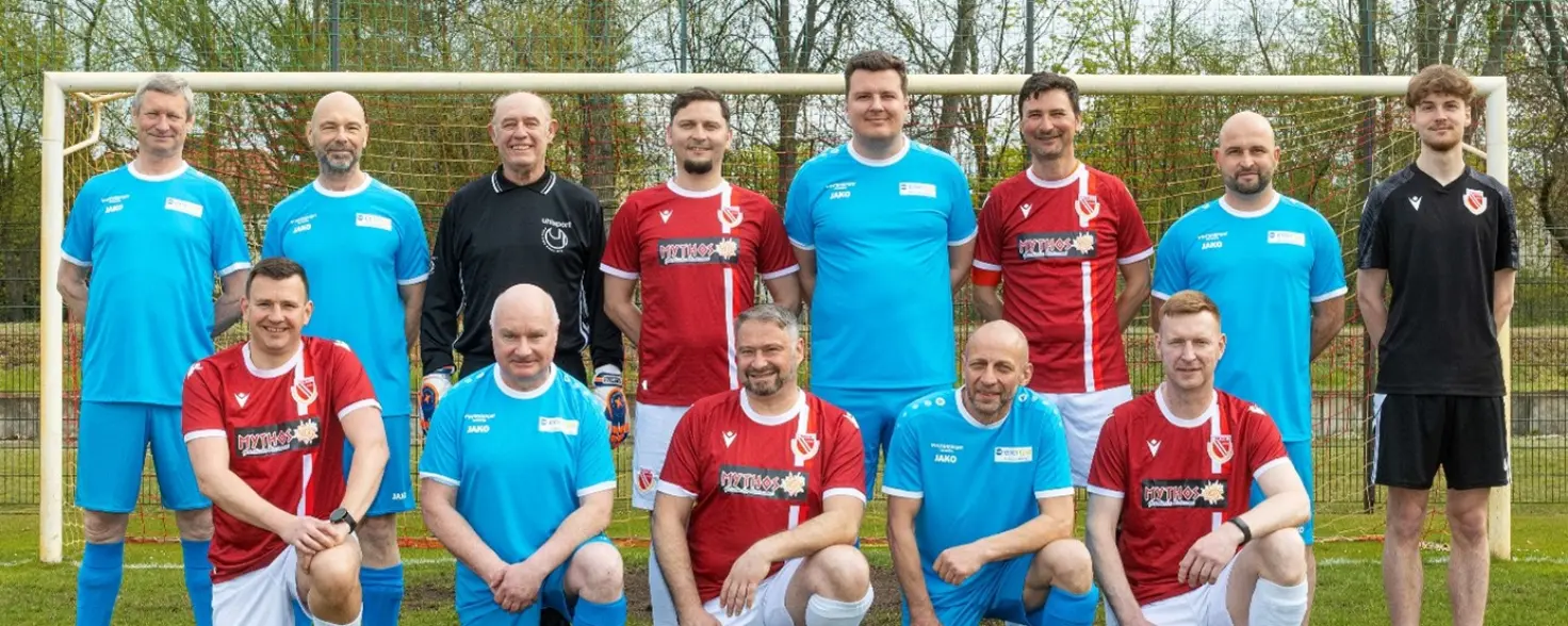 13 Männer in Sportkleidung stehen auf einem Fußballplatz vor einem Tor.