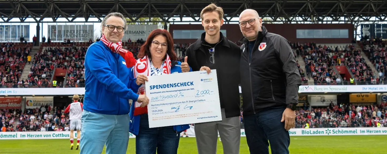Vier Personen stehen nebeneinander in einem Fußballstadion auf dem Rasen. Gemeinsam halten sie einen Spendenscheck im Wert von 2000 Euro.
