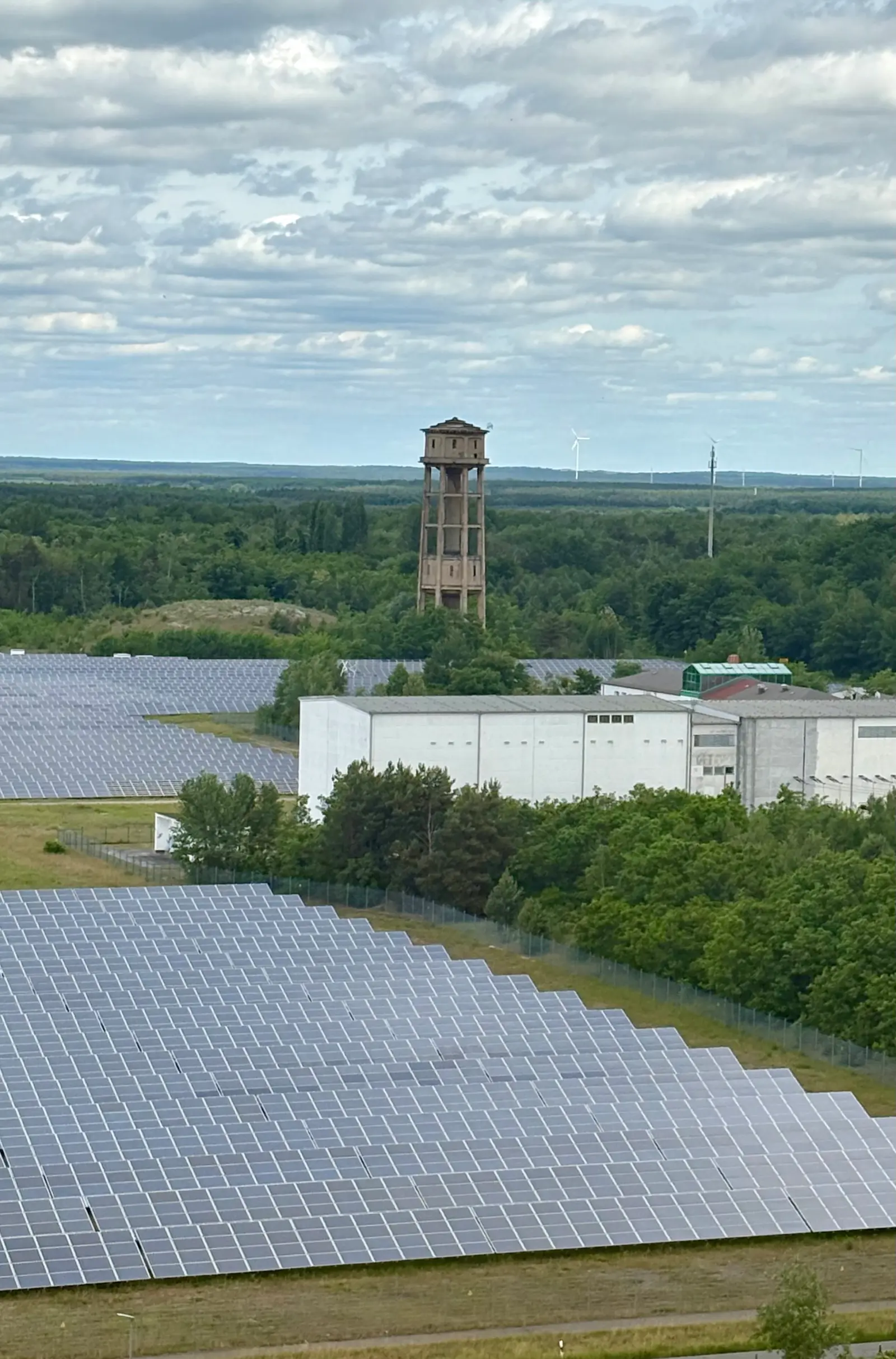 Solarpark mit großflächigen Photovoltaikanlagen, Industriebauten und historischem Wasserturm in bewaldeter Landschaft.