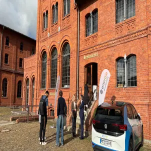 Sechs Menschen stehen vor einem großen Backsteingebäude und steigen die Treppe zum Eingang hoch. Rechts steht ein Elektroauto..