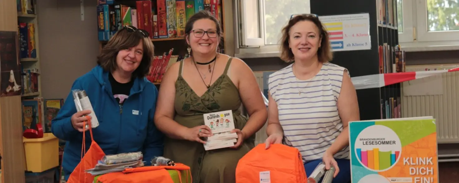 Drei erwachsene Frauen sitzen vor orangenen Turnbeuteln in einer Bibliothek.