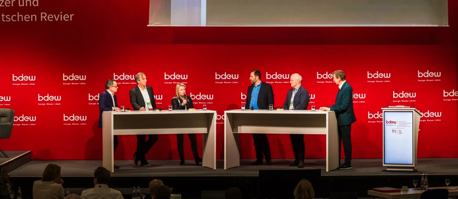 Auf der Bühne findet eine Podiumsdiskussion mit sechs Menschen statt.