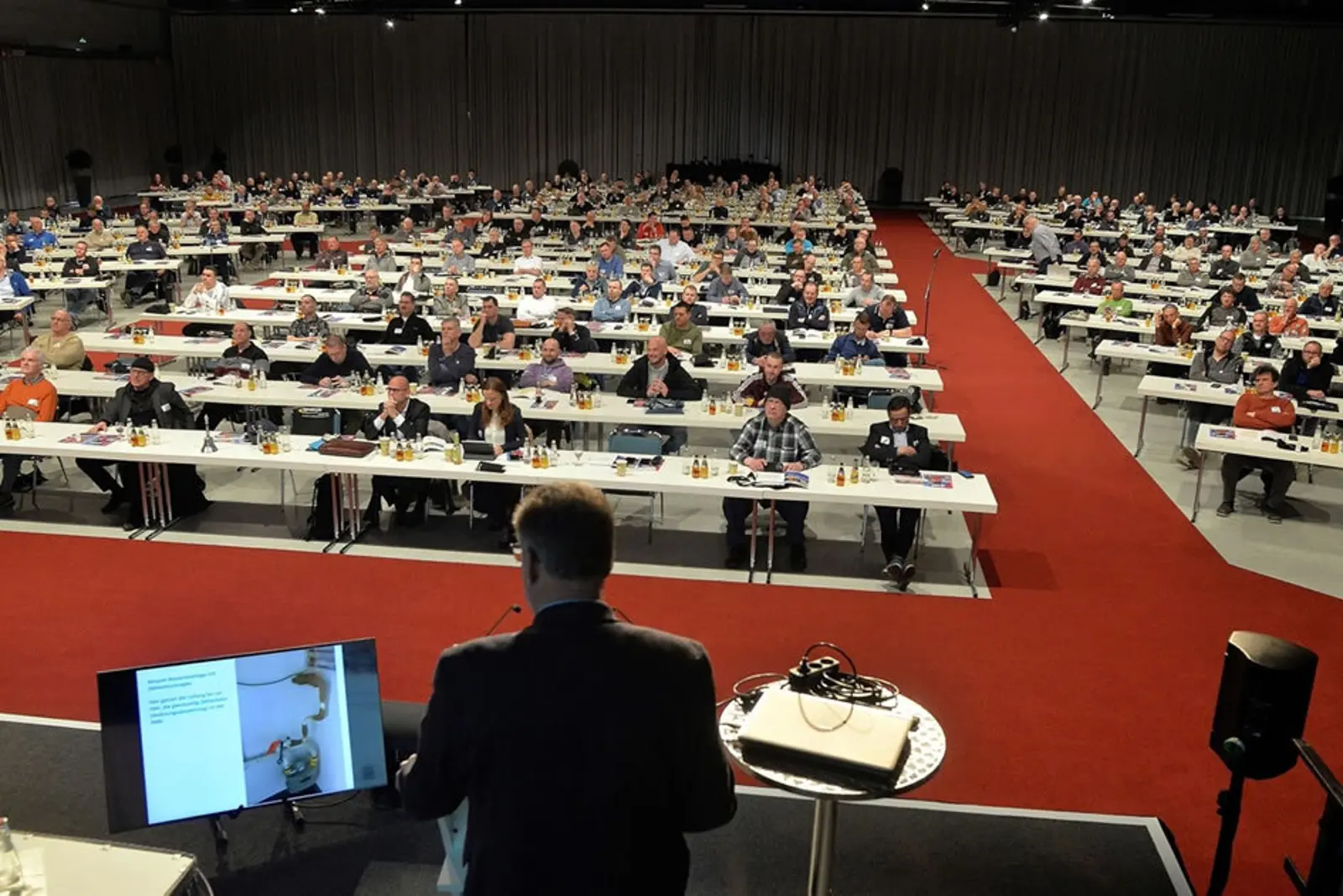 In einem großen Saal sitzen viele Menschen an Tischen mit Laptops und hören den Vorträgen zu