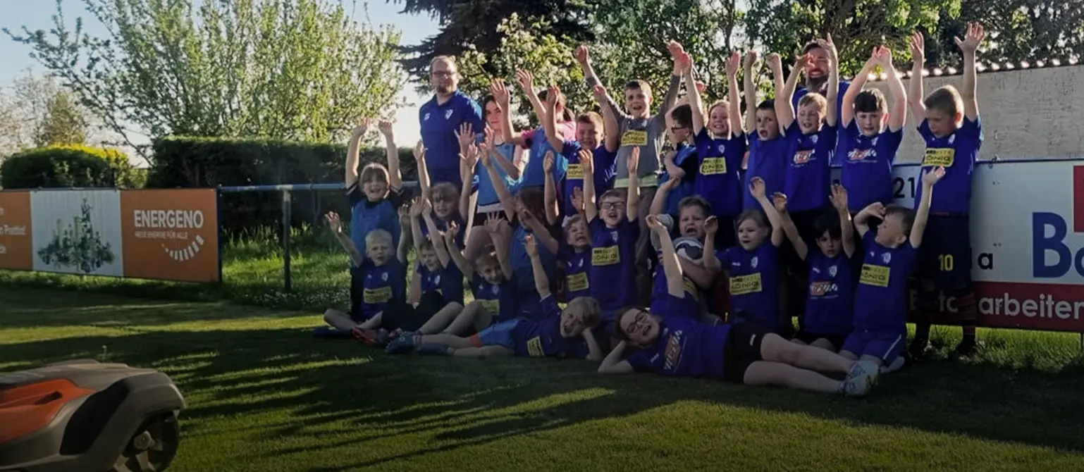 Ein Mähroboter fährt über die große Fußballfläche und die TSV Mannschaft hat sich jubelnd hinter ihm aufgereiht zum Gruppenfoto