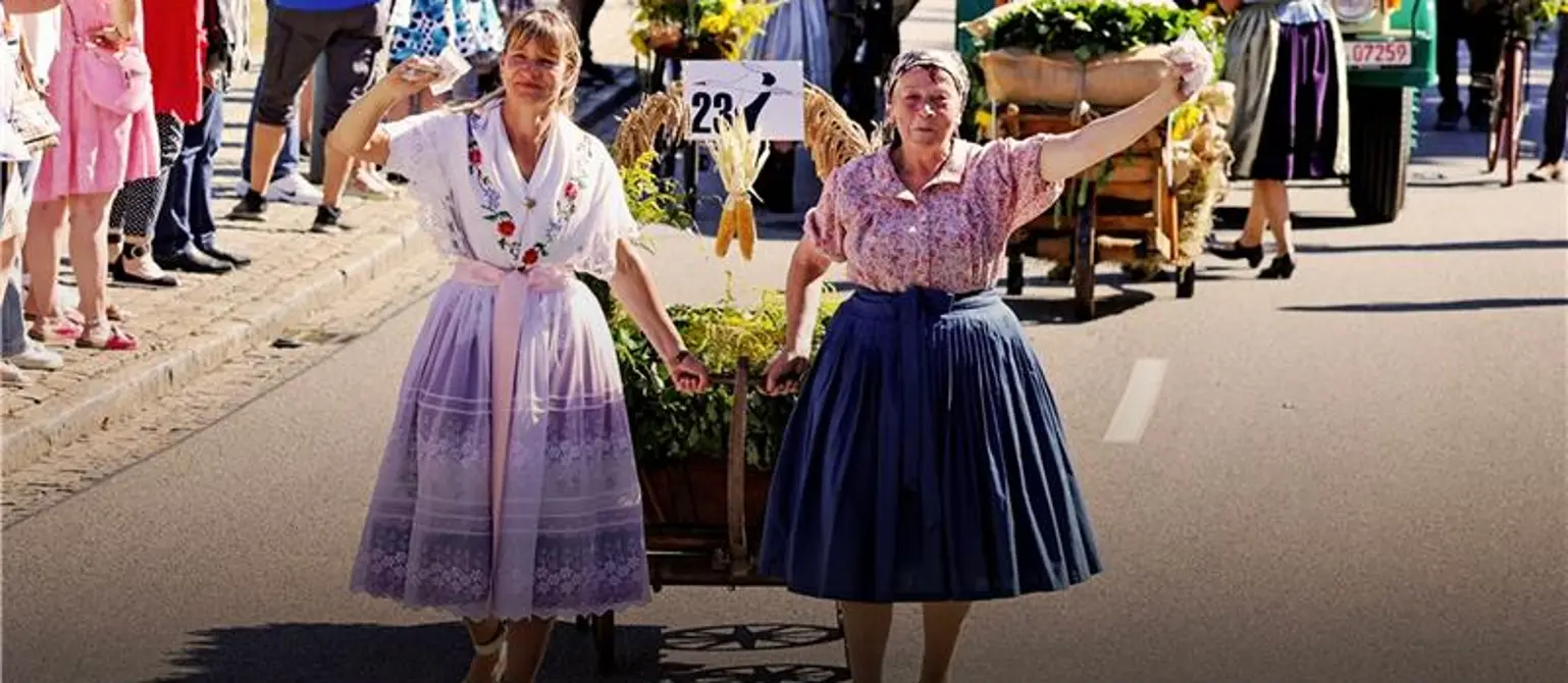 Zwei Frauen in Tracht ziehen beim Einem Umzug im Amt Burg im Spreewald einen Blumenwagen. Zuschauer säumen die Straße, ein grüner Oldtimer-Lkw folgt.