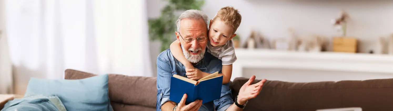 Ein Großvater liest seinem Enkel aus einem Buch vor. Der Enkel umschlingt von hinten seine Schultern.